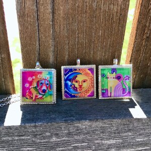 May include: Three silver framed square pendants with colorful images. The first pendant features a dachshund dog with a rainbow background. The second pendant features a sun and moon with a blue and purple background. The third pendant features a cat with a green and purple background.