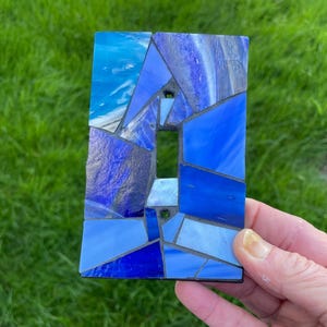 May include: A rectangular light switch cover plate featuring a mosaic design composed of various shades of blue glass pieces. The glass fragments are arranged in an irregular geometric pattern with dark gray grout lines between each piece.