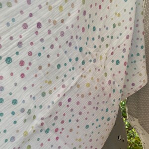 May include: White muslin fabric with a pattern of colorful, smiling face dots in shades of pink, purple, yellow, and teal. The fabric is slightly translucent, with a crinkled texture. Other fabrics are visible in the background.