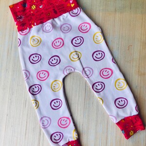 May include: White baby pants with a pink, purple, and yellow smiley face pattern. The pants have a red waistband with a white and pink doodle pattern.