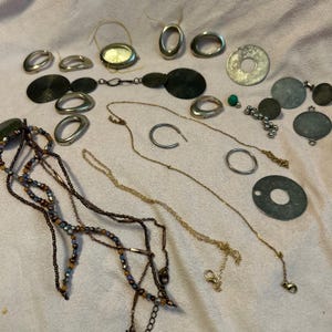 May include: A selection of jewellery items, including necklaces, rings, and pendants, in various metallic tones. The assortment features beaded necklaces in brown and blue hues, along with gold and silver chains and circular elements.