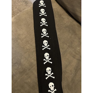 Puede incluir: Tela negra con un patrón repetido de calaveras y huesos blancos. Algunas calaveras tienen parches en los ojos. El diseño es un tema pirata clásico, adecuado para ropa o accesorios.