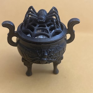 May include: A dark gray metal incense burner shaped like a small cauldron with three legs and two side handles. The lid features a raised spider and web design, while the body is decorated with a relief of a spider crawling on a web pattern.