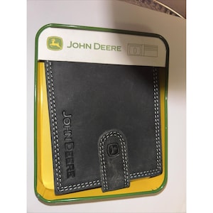 Puede incluir: Cartera de cuero negro con costuras blancas, grabada con el logotipo y el nombre de John Deere. La cartera está en una lata verde y amarilla de la marca John Deere.