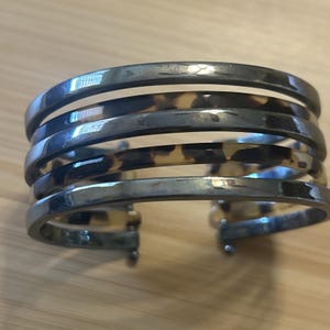 May include: A multi-strand cuff bracelet featuring alternating bands of polished dark metallic silver and a brown and black tortoiseshell pattern. The open-ended design consists of five distinct horizontal rows connected at the sides.