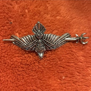 Op de afbeelding: Een zilverkleurige metalen broche in de vorm van een vogel met gespreide vleugels. De vogel is horizontaal geplaatst, met een lange, dunne speld die door het midden van het lichaam loopt. Het metaal heeft gedetailleerde texturen op de vleugels.