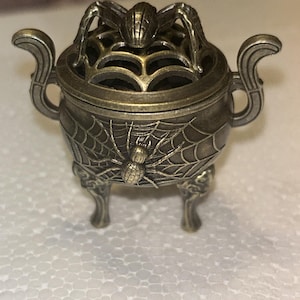 May include: A small, antique-style bronze-toned metal incense burner featuring a three-legged cauldron design. The exterior is decorated with a raised spider and web motif, while the removable lid displays a spider atop a web-patterned grate.