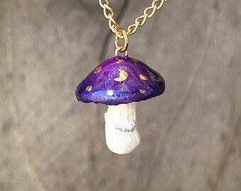 May include: A gold chain necklace with a purple and gold mushroom pendant. The mushroom has a white stem and a purple cap with gold glitter and crescent moon shapes.