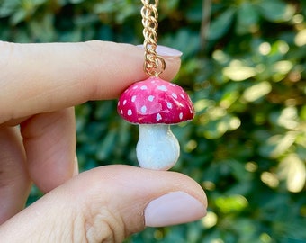 May include: A gold chain necklace with a red and white mushroom pendant. The mushroom has white dots on the red cap and a white stem.