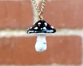 May include: A gold chain necklace with a black and white mushroom pendant. The mushroom has white spots on the top and a white stem.