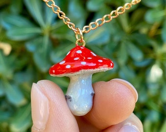 May include: A gold chain necklace with a red and white mushroom pendant. The mushroom has white spots on the red cap and a white stem.