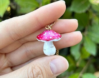May include: A pink and white mushroom pendant necklace with a gold chain. The mushroom has white dots and a crescent moon design on the cap.