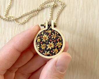 May include: A gold chain necklace with a round wooden pendant featuring a floral embroidery design with yellow and orange flowers on a dark brown background.