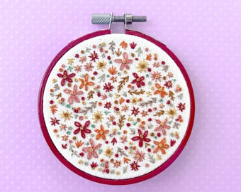 May include: A white embroidery hoop with a red wooden frame, featuring a floral design with red, orange, and yellow flowers and leaves.