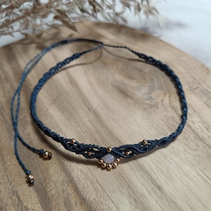 Peut inclure: Un collier ras du cou macramé bleu marine avec un motif central orné d'une petite pierre rose clair et de perles dorées. Le collier est présenté sur une surface en bois, mettant en valeur ses détails faits à la main.