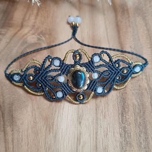 Könnte beinhalten: Ein Makramee-Armband mit einem zentralen ovalen Labradorit-Stein mit blauem Schimmer, umgeben von dunkelgrauem und goldenem Kordelgeflecht. Das Design enthält kleine weiße Perlen und goldene Akzente sowie einen verstellbaren Kordelverschluss.