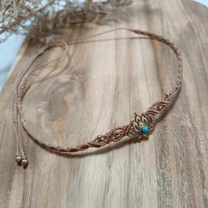 Può includere: Una collana girocollo in macramè fatta a mano nei toni del beige e del marrone. La collana presenta un ciondolo centrale a forma di fiore di loto color oro con una piccola pietra turchese. Piccole perline dorate accentuano il design. La collana è esposta su una superficie di legno.