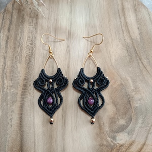 May include: A pair of handmade black macrame earrings with gold-colored hooks and accents. Each earring features a teardrop shape with a central purple bead and a small gold bead at the bottom. The earrings are displayed on a wooden surface.