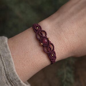 Op de afbeelding: Een handgemaakte macramé armband in een diepe bordeauxrode kleur. De armband heeft een centrale ronde, rood-paarse steen en kleine goudkleurige kralen. De macramé knopen creëren een decoratief patroon. De armband wordt om de pols gedragen.