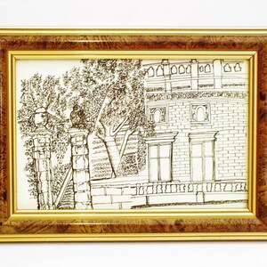 以下が含まれることがあります： 建物と風景の額入りペンとインクの絵。アートワークは、金色の内側の境界線と濃い茶色の木目模様の外側の境界線が付いた長方形のフレームに入っています。この絵は、アーチ型の窓と木のある建物を描いています。