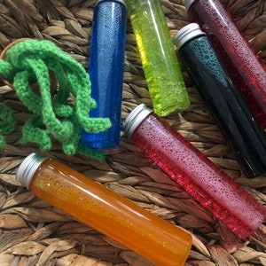 May include: Six glass tubes filled with colorful liquids and glitter, with silver lids. The tubes are arranged in a woven basket.
