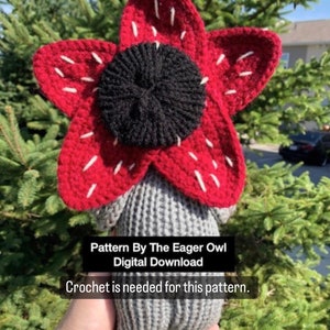 Puede incluir: Un juguete de peluche tejido a crochet con forma de flor roja con un centro negro. El juguete está siendo sostenido en una mano. El texto "Pattern By The Eager Owl Digital Download Crochet is needed for this pattern." es visible debajo del juguete.