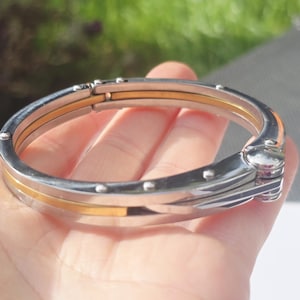 May include: A silver and gold-tone hinged bangle bracelet. The bracelet features a polished silver exterior with a gold-tone center band. Small silver rivets adorn the outer edges. The bracelet is approximately 6.35 cm in diameter.