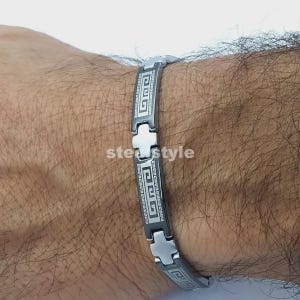 Op de afbeelding: Een zilveren en zwarte armband met een Grieks sleutelontwerp. De armband is gemaakt van metaal en heeft een reeks rechthoekige schakels met een herhalend geometrisch patroon. De tekst "ste style" is zichtbaar op de armband.