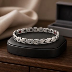 May include: A silver-toned metal link bracelet featuring a repeating leaf-like pattern. The interior of the bracelet displays small, circular embedded accents in red, black, and silver colours along the inner band, secured with a metal clasp.