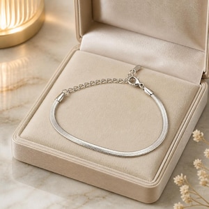 Puede incluir: Una pulsera plateada de espiga exhibida dentro de una caja de joyería beige. La pulsera tiene un diseño plano, similar a una serpiente, y una cadena ajustable. La caja está abierta, revelando un interior suave y aterciopelado, sobre una superficie de mármol.