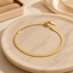 Könnte beinhalten: Ein goldfarbenes Armband mit Fischgrätenmuster. Das Armband wird auf einem weißen Marmorteller präsentiert. Das Armband hat ein rechteckiges Etikett und eine verstellbare Kette. Es ist ein Schmuckstück.