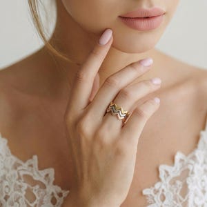 Puede incluir: Una mano que lleva tres anillos apilables con diseño de chevrón en oro, oro rosa y plata. Los anillos tienen un diseño ondulado en zigzag. La persona tiene la piel clara y lleva un top de encaje blanco.