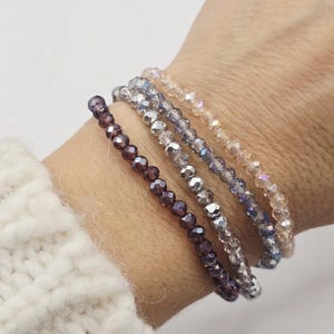 Puede incluir: Cuatro pulseras de cuentas en tonos morado, plateado y transparente. Las pulseras están hechas de pequeñas cuentas facetadas ensartadas. Las pulseras se llevan en la muñeca, con un suéter blanco en el fondo.