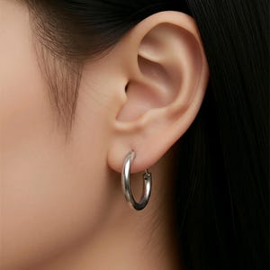 May include: A single silver hoop earring is shown in an ear. The earring has a simple, rounded design and is made of a shiny, metallic material. The earring is a classic style and is suitable for everyday wear.