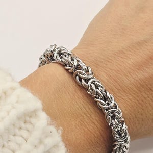 Puede incluir: Un brazalete de cadena bizantina de color plateado. El brazalete está hecho de anillos de metal entrelazados, creando un diseño texturizado y detallado. El brazalete se usa en una muñeca, mostrando su intrincada artesanía y acabado pulido.