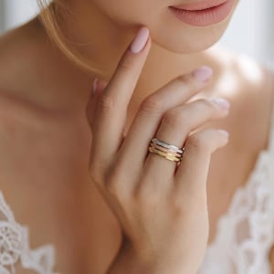 Può includere: Una mano che indossa quattro anelli impilabili strutturati in oro, oro rosa e argento. Gli anelli sono sull'anulare. Lo sfondo è sfocato, con un focus sugli anelli e sulla mano.