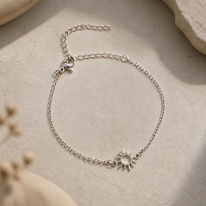 May include: A silver-toned metal chain bracelet featuring a small, circular sun charm with radiating rays. The bracelet includes a delicate link chain, a lobster claw clasp, and an adjustable extension chain with small circular links at the end.