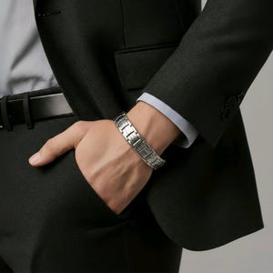May include: A silver bracelet with a rectangular link design. The bracelet is worn on a wrist, with a dark suit jacket and light blue shirt cuff in the background. The bracelet is made of metal and has a polished finish.