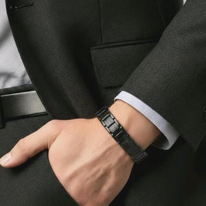 May include: A black rectangular link bracelet is worn on a wrist. The bracelet is made of dark metal and is worn over a light blue collared shirt and a black suit jacket. The person's hand is resting on a black suit.