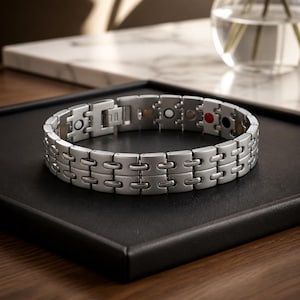 May include: A silver metal bracelet with a rectangular link design. The bracelet has a clasp closure and small coloured accents, including gold, white, red, and black. The bracelet is displayed on a black surface.