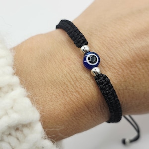 Könnte beinhalten: Schwarz gewebtes Armband mit einem blauen und weißen Evil-Eye-Perle. Das Armband hat einen verstellbaren Knotenverschluss. Die Evil-Eye-Perle befindet sich in der Mitte, flankiert von silberfarbenen Perlen. Das Armband ist verstellbar.