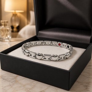 May include: A silver-toned link bracelet featuring alternating heart-shaped charms and H-shaped metal links. The bracelet includes small circular accents in red and black on the inner side of the links, presented inside a black jewelry box.