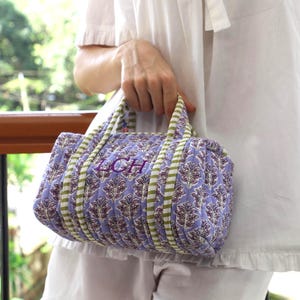 May include: A quilted lavender and brown patterned duffel bag with green and white striped accents. The bag has a top handle and the initials "LCH" embroidered on the side. The bag is approximately 12 inches long.