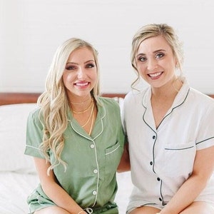 Op de afbeelding: Twee vrouwen in bijpassende pyjama's, een in groen en een in wit, met zwarte bies. Ze zitten op een bed en glimlachen naar de camera.