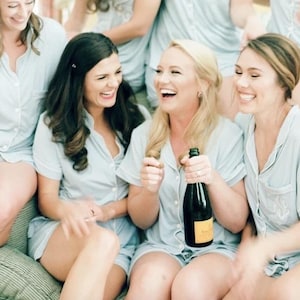 May include: A group of women in light blue pajama sets laughing and smiling. One woman holds a dark bottle with a yellow label. The pajama sets have short sleeves and shorts. The scene suggests a celebratory event.