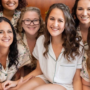 Op de afbeelding: Vijf vrouwen in bijpassende pyjama's met bloemenprint en witte bies. De vrouw in het midden draagt een witte pyjama top met groene bies en een gouden ketting. De vrouwen lachen en kijken naar de camera.