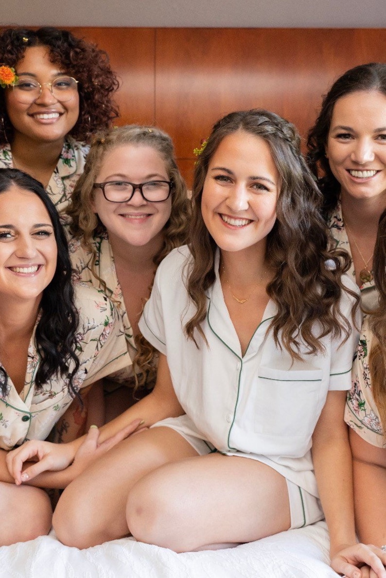 Op de afbeelding: Vijf vrouwen in bijpassende pyjama's met bloemenprint en witte bies. De vrouw in het midden draagt een witte pyjama top met groene bies en een gouden ketting. De vrouwen lachen en kijken naar de camera.