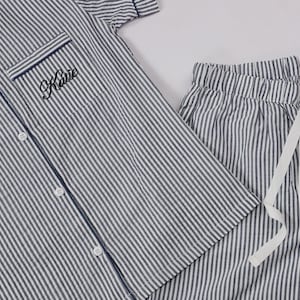 May include: A short-sleeved button-down pajama shirt and matching drawstring pajama pants featuring a navy blue and white vertical stripe pattern. The shirt includes a chest pocket embroidered with the name Katie in black cursive script lettering.