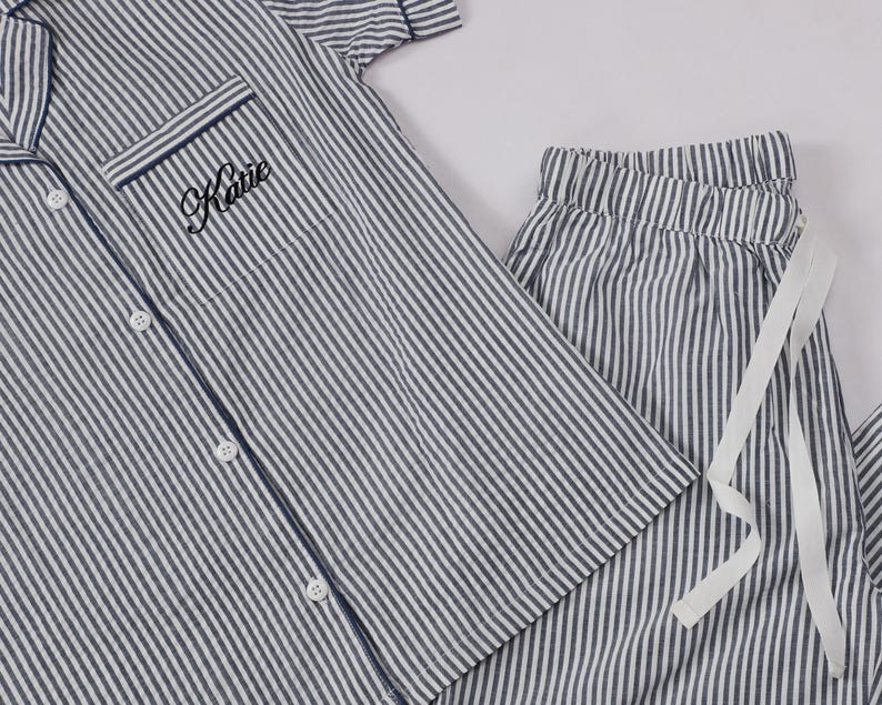 May include: A short-sleeved button-down pajama shirt and matching drawstring pajama pants featuring a navy blue and white vertical stripe pattern. The shirt includes a chest pocket embroidered with the name Katie in black cursive script lettering.