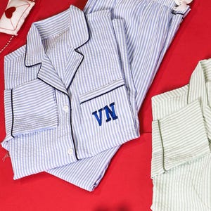 May include: A set of blue and white striped pajamas featuring a button-down shirt with a collar, long sleeves, and the letters VN embroidered in blue on the chest pocket. The matching pants have an elastic waistband with a white ribbon tie.